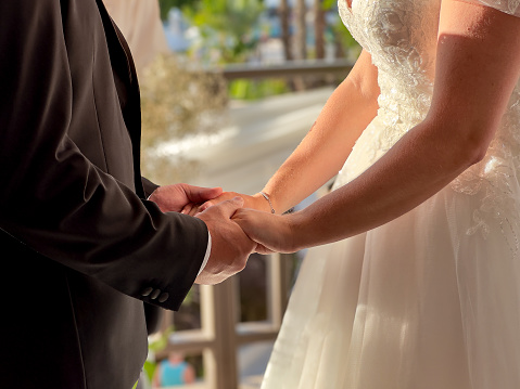 Close-up of groom and bride in black suit holding bride's hands on wedding ceremony day.