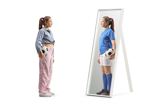 Full length shot of a girl holding a football and looking at a mirror at home