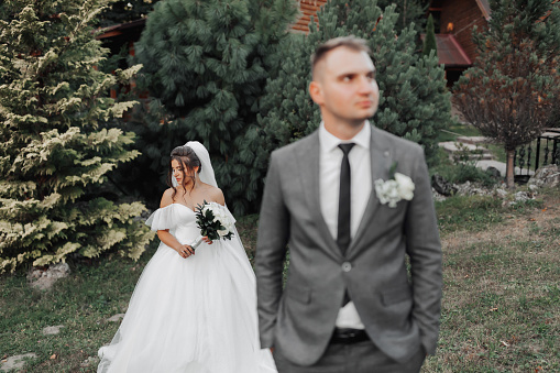 portrait of the bride and groom in nature. The groom is in the foreground, blurred, the bride stands behind him in a white voluminous dress, holding a bouquet and looking down. Stylish groom