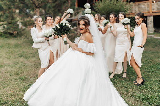 Wedding photo in nature. A brunette bride in a long white dress stands in front of her friends in nude dresses, who stand smiling, holding up their gypsophila bouquets. Young women. emotions