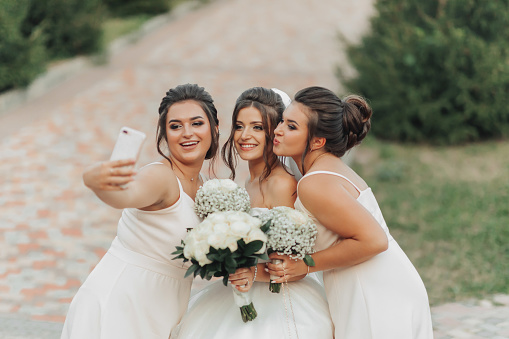 Wedding photo in nature. A brunette bride in a long white dress and her friends in nude dresses are smiling and taking a selfie on their phone, holding their gypsophila bouquets. Young women. emotions