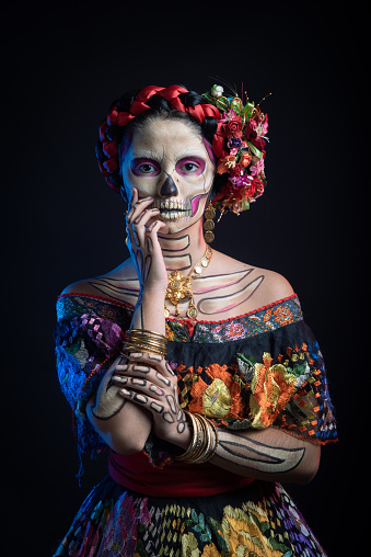 Latina woman with vibrant skull makeup, dressed in a Chiapas floral embroidered dress, against a dark backdrop.