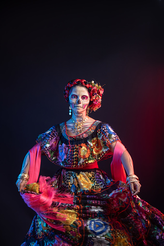 Woman of Latin descent in a Catrina costume, displaying elaborate skull makeup and a Chiapas floral embroidered dress.