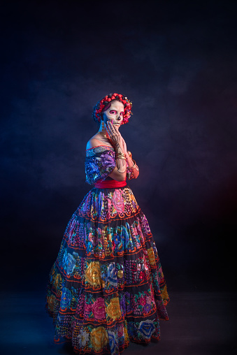 Latina lady dressed as a Mexican Catrina, with intricate skull makeup and a Chiapas floral embroidered dress, on a dark backdrop.
