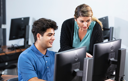 A high school student and a teacher in a computer lab. The student, a Middle Eastern teenage boy, is using a desktop PC, and the teacher, a young woman in her 20s, is standing next to him pointing at the screen.
