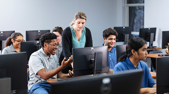 A multiracial group of high school students in a computer lab, each using a desktop PC. The teacher is helping one of the students, a black teenage boy.