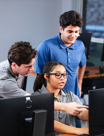 A multiracial group of three high school students in a computer lab working on a project together. A mixed race teenage girl is sitting at a desktop PC, between two male classmates. They are all 17 years old.