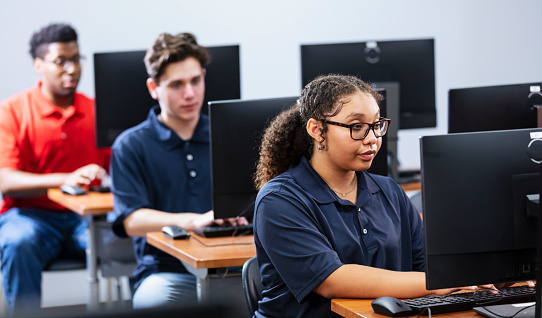 A multiracial teenage girl in a high school computer class with two teenage boys using desktop pcs.