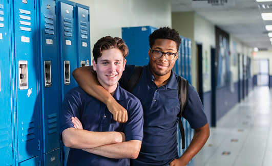 Two multiracial high school students standing together in the school hallway next to their lockers. The teenage boys are smiling confidently at the camera. The African-American student is 18 years old, and his friend is 17.