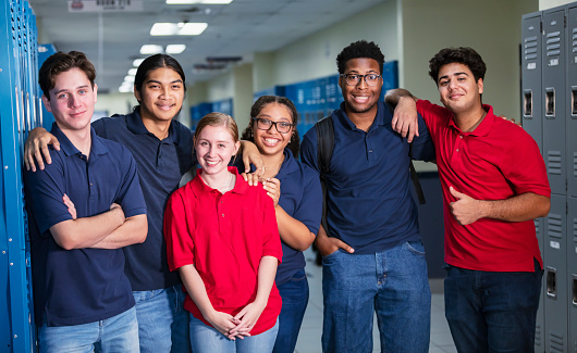 A multiracial group of six high school students standing together in the school hallway smiling and looking at the camera.