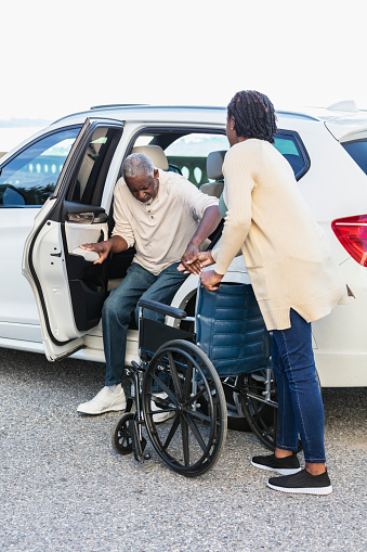 A senior African-American man getting into or out of a car with help from his adult daughter, a mature woman in her 40s, who is standing behind the senior's wheelchair, holding his hand for support.