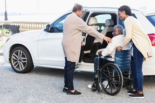 A senior African-American man in a wheelchair, traveling by car. He, his wife, and his daughter, are next to the open door of the car.