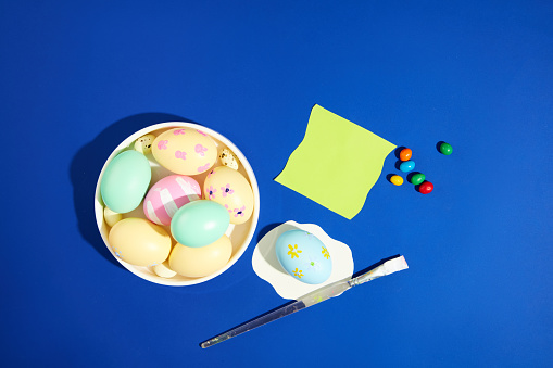 Happy Easter Day concept with a white dish containing many Easter egg painted in pastel colors arranged with colorful candies and a brush. A note paper with copy space for Easter content