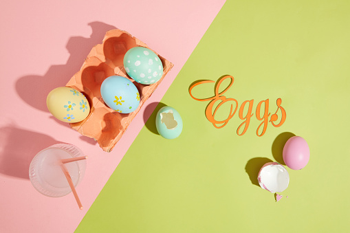 Some lovely Easter eggs contained inside an egg carton painted in coral color. A glass cup filled with water containing two paintbrushes. The word Eggs made of paper featured