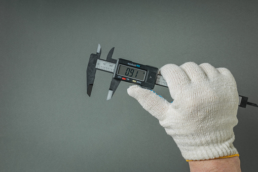 A gloved hand with an electronic caliper on a gray background. A tool for accurate measurement of dimensions.