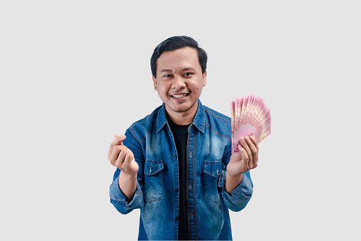 Young Asian man giving love symbol while holding paper money and showing happy expression
