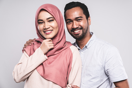 Young happy Malaysian couple standing together side by side, looking towards the camera with a bright happy smile. Malaysian woman with typical modern headscarf, young man in button down shirt.. White Background Portrait Studio Shot. Kuala Lumpur, Malaysia.