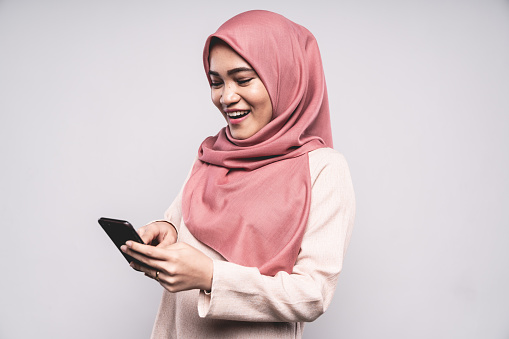 Happy smiling Malaysian Woman standing in Studio, looking down to her Smart Phone with a bright happy natural laugh - smile,  wearing a fashionable modern Hijab - Headscarf. Using Smart Phone - Mobile Apps Concept, White Background Portrait Studio Shot. Kuala Lumpur, Malaysia.