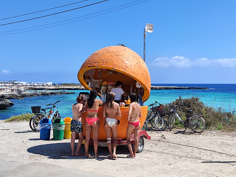 Group of youngsters at an orange shaped juice kiosk by the sea on the island of Favignana in the Egadi Archipelago, Sicily, Italy.