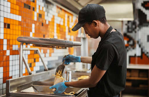 Black man, fries and fast food kitchen worker at fryer with gloves, cooking and order process at small business. Restaurant chain, working and cook preparing chips for lunch service with hospitality.