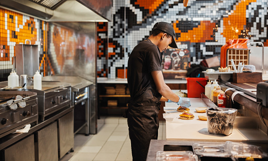 Man, burger and fast food kitchen for prep at counter with gloves, cooking and uniform at small business. Restaurant, sandwich and cook with hamburger patty stack for lunch service with take away.