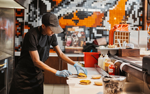 Man, burger and fast food kitchen worker at counter with gloves, cooking and uniform at small business. Restaurant, sandwich and cook with hamburger patty stack for lunch service with take away.