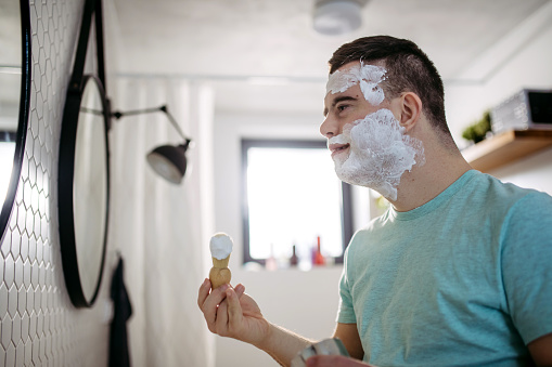 Young man with down syndrome learning how to shave, applying shaving foam, cream all over his face, looking at mirror.