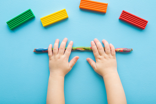 Baby boy hands rolling colorful stick from modeling clay on light blue table background. Pastel color. Closeup. Point of view shot. Toddler development. Making roll shape. Top down view.