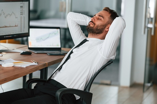 A stock photo of a young businessman sleeping in his office cubicle with his feet kicked up on the desk.