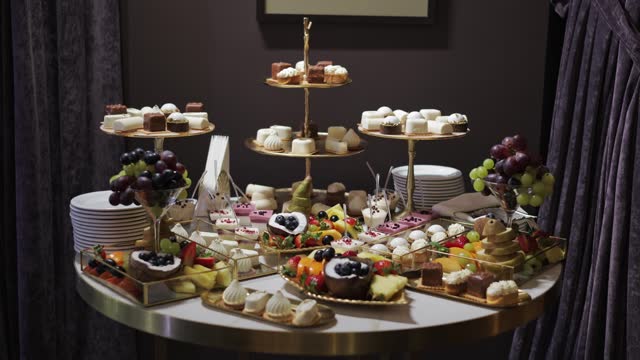 Variety of multiple tiers of dessert trays holding assortment of sweet treats, tarts adorned with fresh berries and other baked goods arranged on round table. Atmosphere of festive catering.