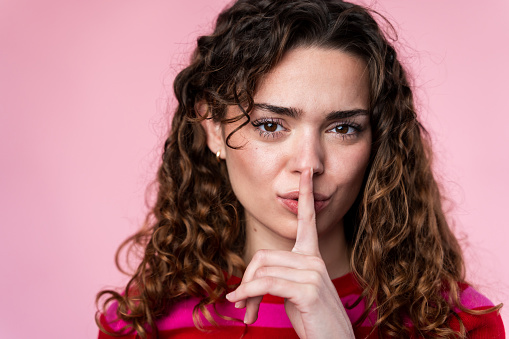 Curly-haired woman in pink putting finger to lips in a shushing gesture