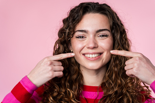 Woman with curly hair cheerfully pointing to her radiant smile, pink background