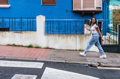 A young woman strides confidently past a vivid blue house, her braided hair flowing as she moves.