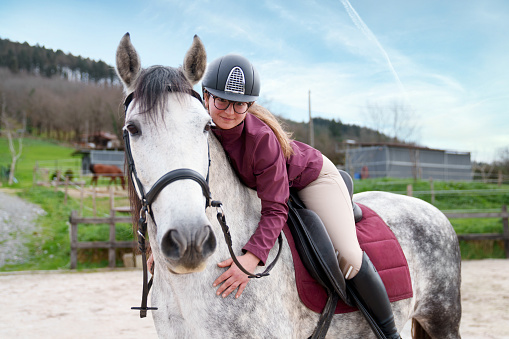 A young rider shares a tender moment with her grey horse during a break in the sandy arena.