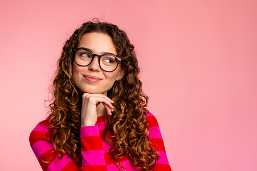 Thoughtful young woman with glasses looking up on a pink background.