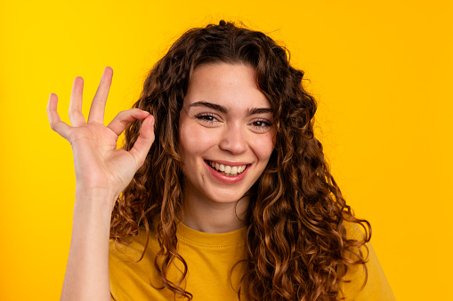 Smiling curly-haired woman giving OK sign with her hand on yellow backdrop