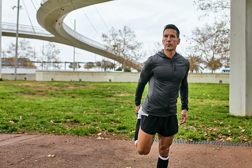 Front view of mature man in sports clothing standing outdoors, preparing for afternoon workout, looking away, footbridge in background.