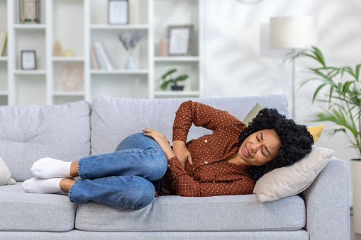 A young African American woman is lying on the sofa at home, holding her stomach with her hands, suffering from severe pain, needing medical treatment.