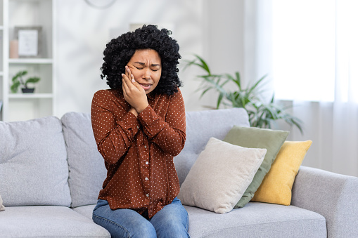 Upset young African American woman is at home, sitting on the couch and closed her eyes, holding her cheek with her hands, having a severe toothache, needing the help of a dentist.