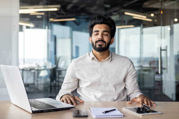 Relaxed young Indian man sitting in office at desk with laptop and documents and resting with closed eyes