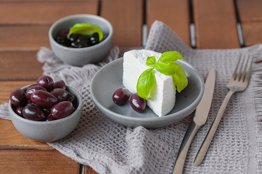 Feta cheese sliced on gray plate with black olives and basil leaves. Delicious and healthy breakfast on terrace