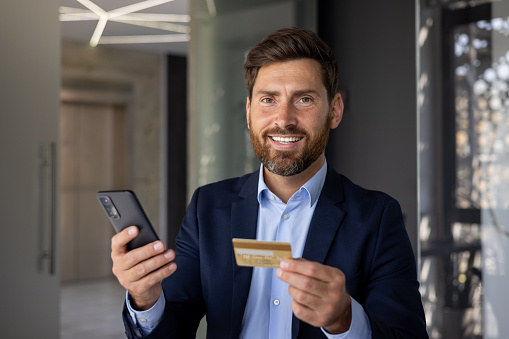 Smiling and successful young man businessman and banker standing in office, holding phone and credit card, looking confidently at camera. Close-up photo.