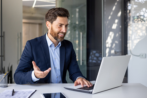 Successful and cheerful young businessman in a suit sitting in the office at the table with a laptop and communicating on a video call gesturing with his hand.