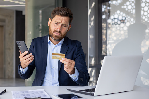 Serious young male businessman sitting in office at desk with laptop and worriedly looking at credit card, holding phone, having financial and technical difficulties.