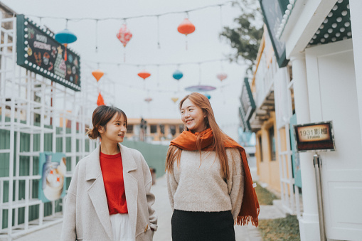 Two young Asian women walking in Hong Kong, chatting outdoors in  Cheung Chau. They are dressed in cozy, autumn clothing with one wearing a beige coat over a red top and the other in a beige sweater with a rust-colored scarf. Behind them, a series of colorful lanterns across the scene, during Chinese New Year celebration.