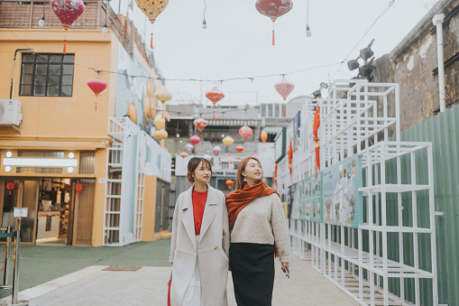 Two asian young women  walking side by side in the streets of Cheung Chau, HongKong during Chinese New Year Celebrations.  The women are dressed in stylish, contemporary clothing. They are looking upwards, likely admiring the lanterns.