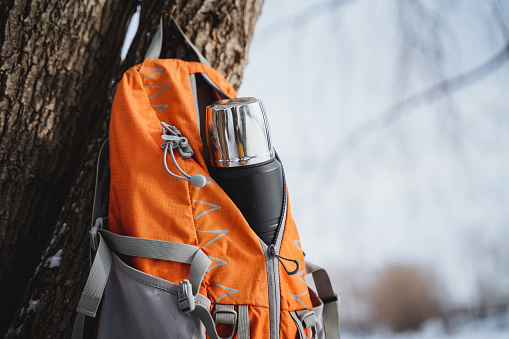 Bright orange and gray backpack with a thermos bottle in the side pocket hangs on a tree. The shiny silver thermos bottle is visible inside. Light brown bark tree in snowy forest.