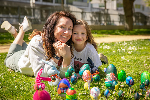 Portrait of a young beautiful mother\nand daughters on a sunny spring day in the park. A woman and a girl are lying on the green grass of the lawn. They hug and smile. In front of them are drying Easter eggs and bunnies, which they decorated with paints and made with their own hands.