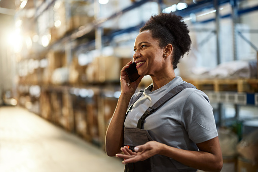 Happy African American female worker communicating over smart phone in a storage room.