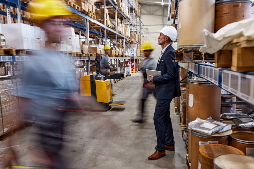 Senior foreman brainstorming while standing in distribution warehouse among workers in blurred motion.